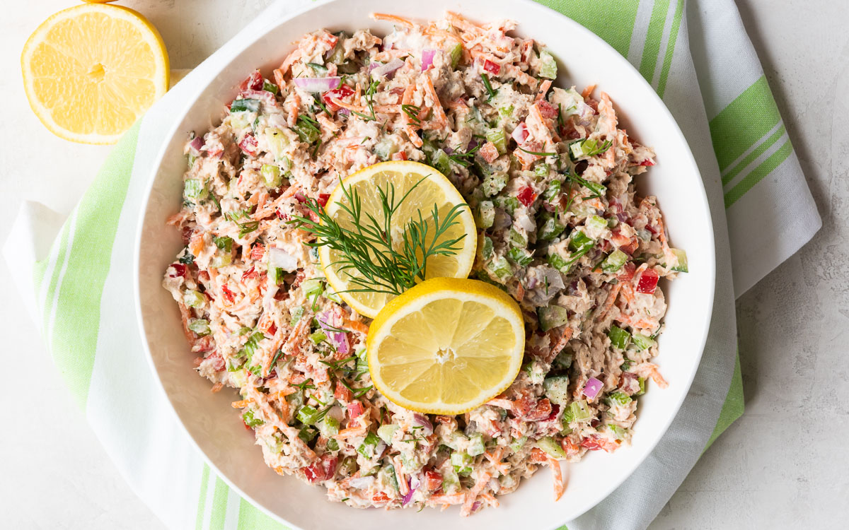 Large overhead photograph of white bowl of salmon salad and chopped vegetables with lemons and dill on top 