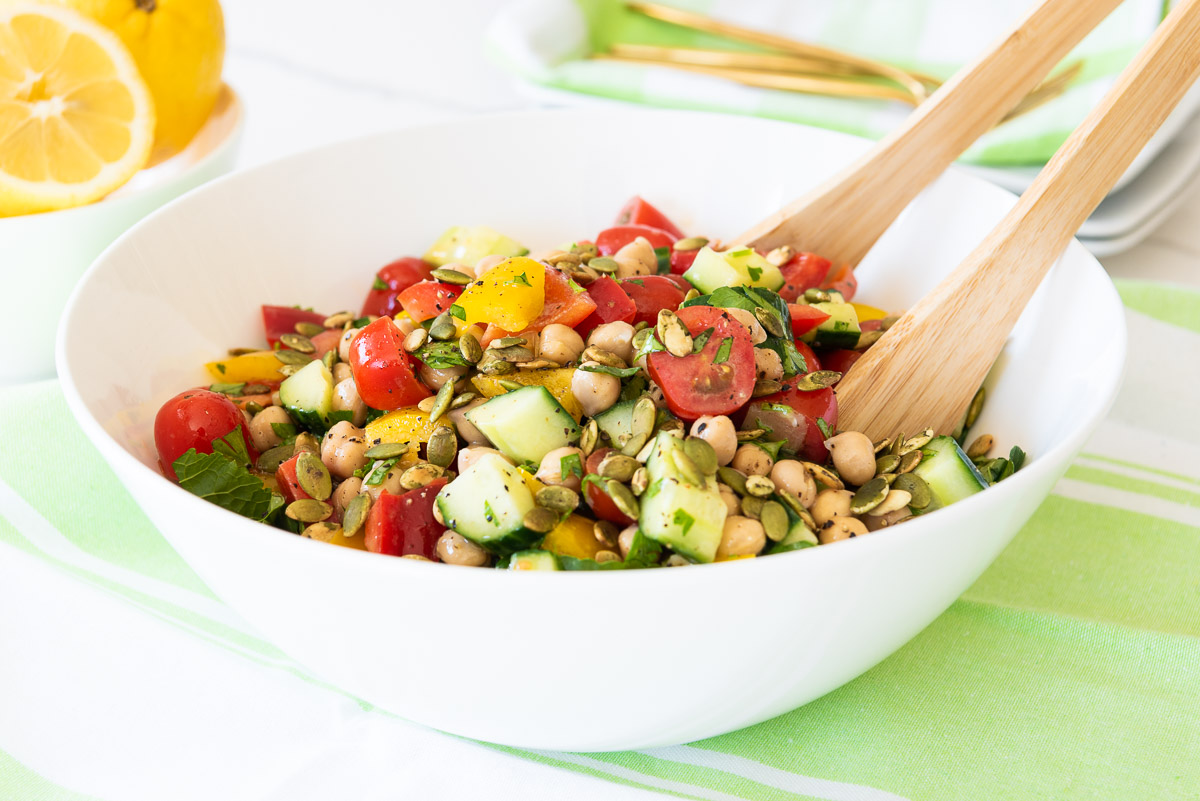 Chickpeas, cucumbers, pepitas, herbs and tomatoes in a large white salad bowl with bright yellow lemons!
